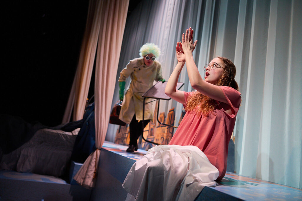 A woman in a pink nightgown sits on the edge of a platform, playing with her hands above her head. She has a sheet across her lap. Behind her, a crazy looking figure in a lab coat with wild blue hair sneaks into the room.