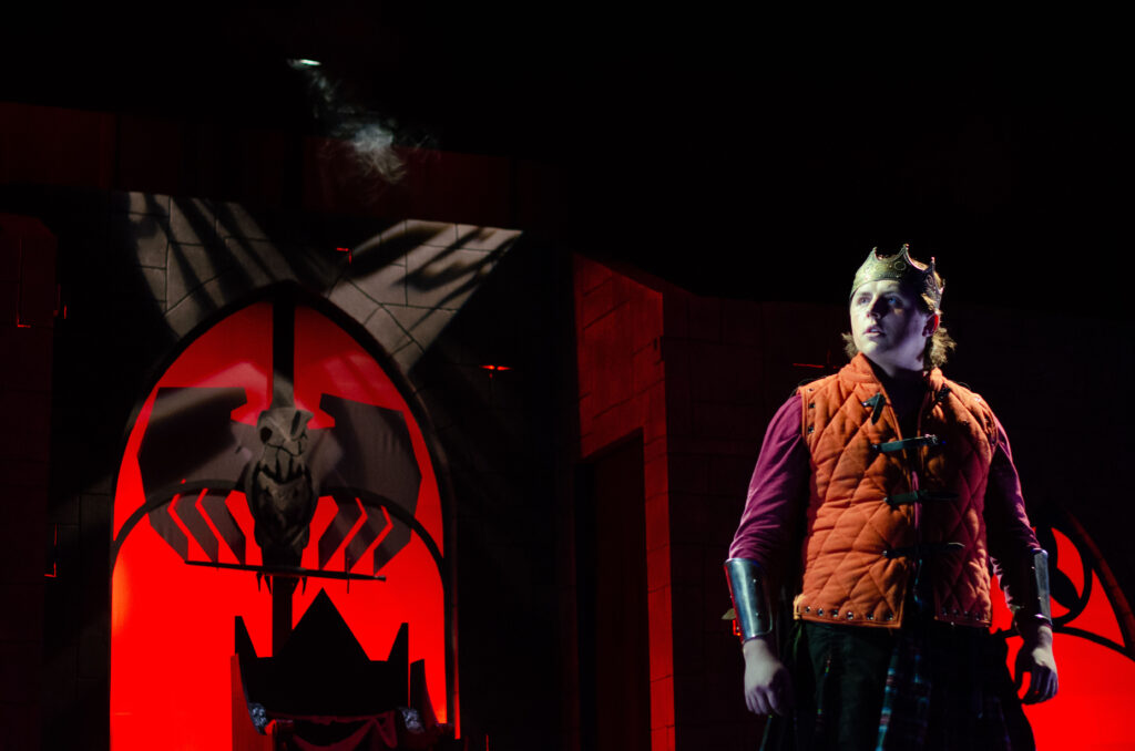 Macbeth stares into the distance, stone faced as he prepares for battle. A large fascist eagle stretches out geometric wings in the window above his throne. The light is all red.