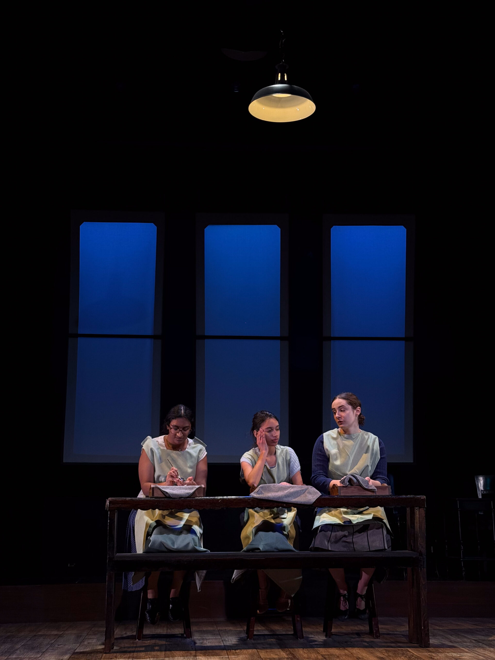 Three young women in aprons sit at an industrial bench chatting and painting numbers on watches.