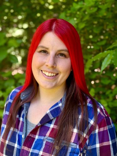 A head shot of Kayleigha. She has long brunette hair streaked with red in the front and smiles for the camera.